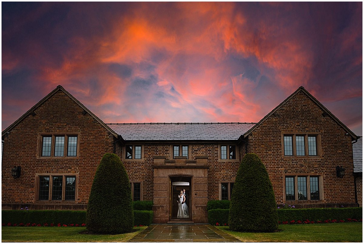 A bride and groom embrace in the doorway of a brick manor against a vibrant sunset sky.